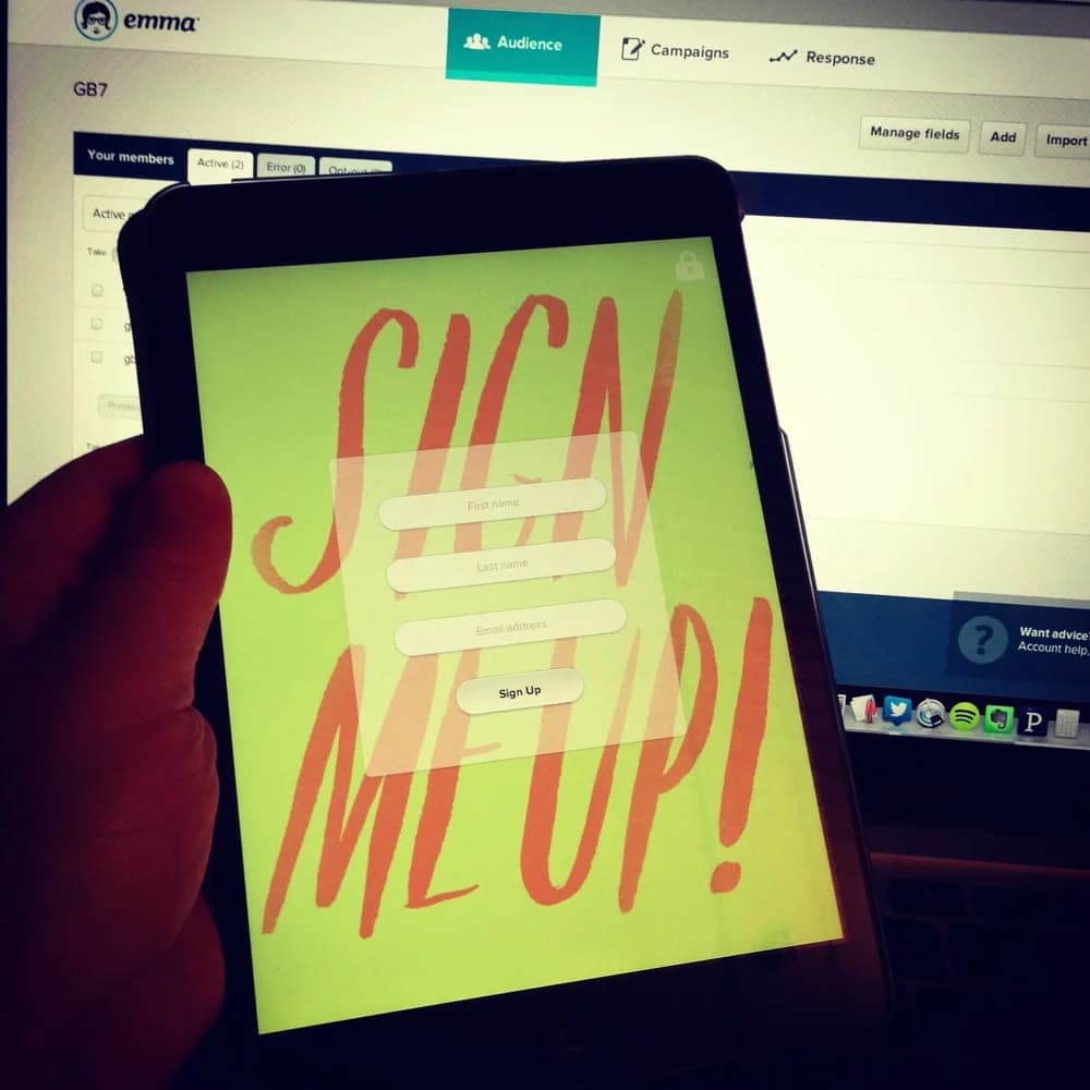 Guestbook running on an iPad Mini held in front of an Emma desktop screen, showing a 'SIGN ME UP!' email signup form.